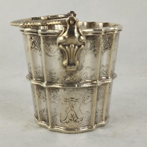Pode incluir: Um pequeno balde de metal prateado com um design de nervuras verticais e padrões intrincados de flores e volutas em relevo. O recipiente inclui uma alça articulada fixada a suportes decorativos nos lados opostos da borda.