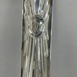 May include: A polished silver-toned metal handle featuring a vertical ray pattern radiating from a central circular emblem. The emblem contains a stylized, embossed design resembling a harp or musical instrument set against a smooth, reflective surface.