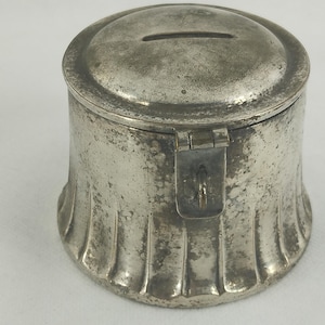 May include: A vintage silver-toned metal bank with a slot on the lid for inserting coins. The bank has a cylindrical body with a flared, ribbed base and a hinged lid secured by a clasp. The metal shows signs of age and wear.