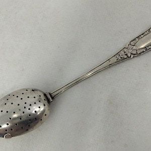 Könnte beinhalten: Ein silberfarbener Teesieb-Löffel aus Metall mit einer ovalen Laffe, die zahlreiche kleine Perforationen aufweist. Der lange Stiel ist mit einem geprägten Blumenmuster und einem glatten Mittelteil verziert und hat ein metallisches Finish.