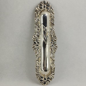 May include: A silver mezuzah case with an elaborate floral and scroll design. The case is rectangular with rounded ends and a central, elongated compartment. The decorative border features detailed carvings.