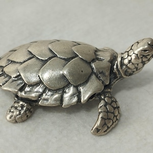 Puede incluir: Una pequeña figura de metal en tono plateado de una tortuga marina. La tortuga presenta un patrón detallado de escamas superpuestas en su caparazón y piel texturizada en la cabeza y las aletas. La figura se muestra de perfil con la cabeza alzada.
