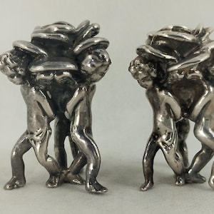 Könnte beinhalten: Zwei silberfarbene Skulpturen, jede mit drei cherubartigen Figuren, die eine große, stilisierte Rose tragen. Die Figuren sind detailliert mit sichtbaren Gesichtszügen und bilden die Basis der rosenförmigen Oberseite. Die Skulpturen werden vor einem neutralen Hintergrund präsentiert.