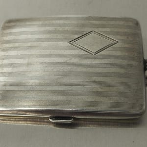 May include: A rectangular silver case with horizontal lines and a diamond-shaped design in the center. The case has a clasp closure and a small loop on the side. The case is likely used for carrying cards or small items.