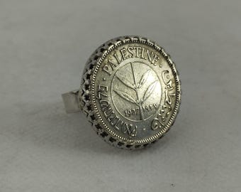 May include: A silver ring featuring a large, round face with the word "PALESTINE" and the year "1927" inscribed. The ring has a detailed, openwork design around the face and a simple band.