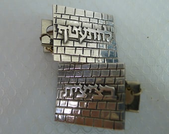 May include: Silver bracelet featuring a brickwork design with Hebrew lettering. The bracelet is made of two rectangular sections joined by a hinge and a small chain. The Hebrew text is raised, creating a textured effect.