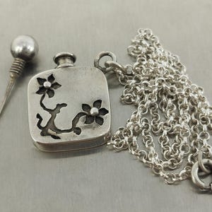 May include: A silver-toned pendant shaped like a small square bottle with a floral cutout design, accompanied by a matching silver-toned chain and a separate spherical-topped stopper. The metal surface has a matte, slightly oxidised finish.