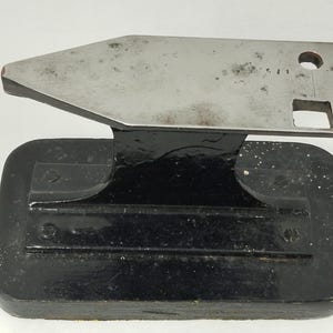 May include: A vintage metal anvil with a black base and a silver top. The anvil has a rectangular base with rounded corners and a flat, angled top. The top has a hole and a square cut-out. The anvil is likely used for shaping metal.
