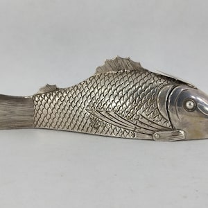 May include: A silver fish-shaped ornament with intricate scale detailing, fins, and a tail. The fish features a hinged lid, possibly for storage or decoration. The item is presented on a simple, neutral backdrop.