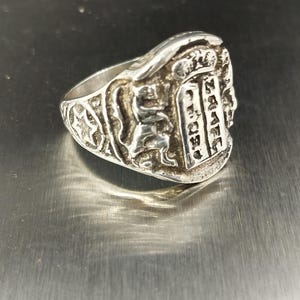 May include: A silver-toned ring with detailed relief carvings. The ring features a central rectangular design with vertical text, flanked by a lion, a star, and other ornate patterns. The ring is set against a metallic surface.