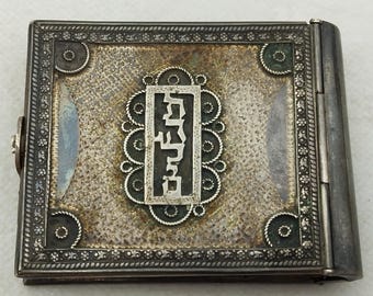 May include: A silver-coloured hinged box with intricate floral designs and Hebrew lettering on the front. The box is closed.