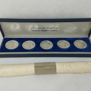 Op de afbeelding: Een set van vijf zilverkleurige ronde munten in een rechthoekige marineblauwe doos met een blauwe fluwelen binnenkant. Het deksel bevat de tekst "Pidyon HaBen Coins" en Hebreeuws schrift. Een opgerolde perkamentrol met een papieren band is inbegrepen.