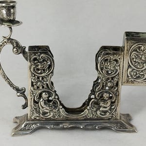May include: A silver-colored metal match holder featuring an attached candle holder on one side. The piece is decorated with intricate embossed floral patterns and small cherub figures on both sides, resting on a wide, ornate rectangular base.