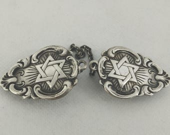 May include: Two silver clasps linked by a chain. Each clasp has a Star of David design in the centre, framed by decorative scrollwork. The clasps are likely for a garment or accessory.
