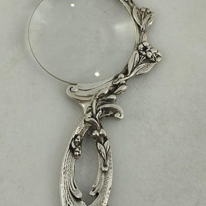 May include: A silver-toned magnifying glass with a clear lens and an ornate handle. The handle and frame are decorated with floral and leaf motifs. The magnifying glass is designed with a decorative, vintage style.
