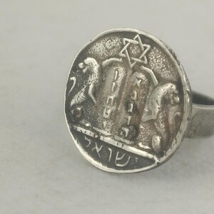 May include: A silver-toned ring featuring a detailed coin design. The coin depicts a Star of David, Hebrew text, and other symbolic imagery. The ring has a simple band and a circular face, showcasing the coin's intricate details.
