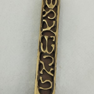 Op de afbeelding: Een rechthoekig metalen object in messingkleur met het woord "YESSE" uitgesneden in een decoratief lettertype. De achtergrond is donkerbruin. Het object heeft twee gaten aan de boven- en onderkant.