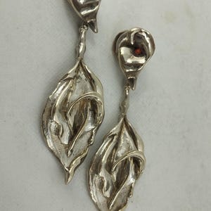 May include: A pair of silver-toned earrings featuring a calla lily design. Each earring has a flower bud with a small red accent and a long, leaf-shaped pendant. The earrings are detailed with textured surfaces and are likely made of metal.