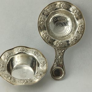 May include: A silver tea strainer with a floral design and a matching silver bowl. The strainer has a handle and a perforated bottom for straining tea leaves.