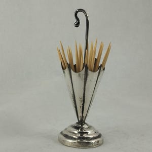 Puede incluir: Un palillero plateado con forma de paraguas. El soporte tiene un asa curvada y un cuerpo acampanado y acanalado. Está lleno de palillos de madera. La base es redonda y tiene un borde decorativo.