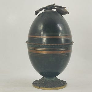 Puede incluir: Un recipiente antiguo en forma de huevo con una pátina verde oscuro. Presenta bandas de color cobre y un elemento decorativo en la parte superior. El recipiente se asienta sobre una base detallada y ornamentada con un borde dorado.