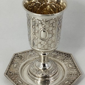 Può includere: Un calice color argento con intricati disegni a griglia e ovali, appoggiato su un piatto esagonale abbinato. Il calice presenta un bordo dettagliato e una base decorativa, che mostra uno stile classico e ornato.