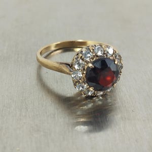 Può includere: Un anello in oro con una grande pietra centrale rotonda di colore rosso intenso, circondata da un alone di piccole pietre chiare e scintillanti. Il gambo è liscio e metallico, con una classica montatura circolare che sostiene la pietra.