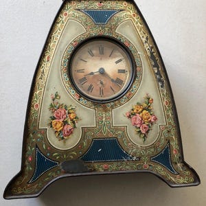 Puede incluir: Reloj antiguo con un diseño floral decorativo en tonos amarillos, rosas y verdes. El reloj tiene una esfera redonda con números romanos y un marco de metal negro.