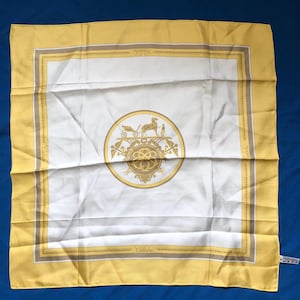 May include: Square silk scarf with a yellow border and a white center. The center features a gold emblem with a horse-drawn carriage and the word "Calèche". The scarf has a gold and brown border.