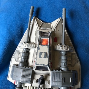 以下が含まれることがあります： スターウォーズのスノースピーダーのヴィンテージおもちゃモデル。車両は主に白で、グレーと赤のアクセントがあり、2つの目立つグレーの大砲と詳細な機械部品が特徴です。おもちゃには使用感が見られます。