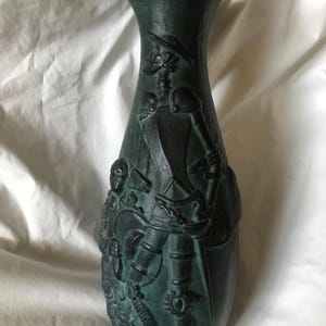 May include: A dark green vase with a detailed relief of a historical figure in armour. The vase has a flared top and a rounded body. The relief depicts a man in a hat and armour, with other figures and decorative elements.
