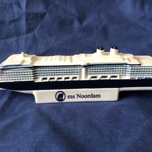 Puede incluir: Un modelo de cerámica blanco y azul del crucero ms Noordam. El barco tiene un casco negro y cubiertas blancas con un patrón azul y blanco. El barco tiene dos chimeneas negras y el texto "ms Noordam" está impreso en la base.