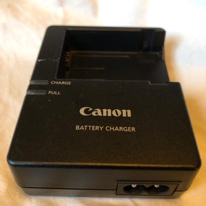 Könnte beinhalten: Ein schwarzes Canon-Batterieladegerät mit dem Text "CHARGE" und "FULL" auf der Vorderseite. Das Ladegerät hat einen Stecker mit zwei Stiften und der Text "BATTERY CHARGER" ist auf der Vorderseite aufgedruckt.