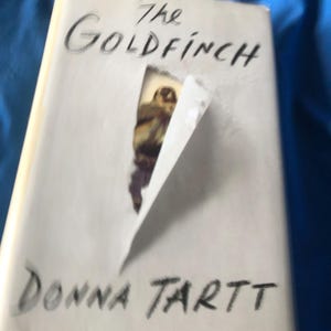 Może przedstawiać: Książka zatytułowana "The Goldfinch" autorstwa Donny Tartt. Okładka jest biała z tytułem i nazwiskiem autora w kolorze czarnym. Rozerwany fragment odsłania częściowy obraz ptaka.