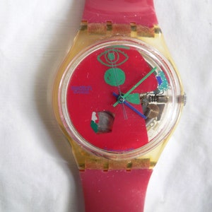 May include: A red, yellow, and green Swatch watch with a clear plastic case and a red silicone band. The watch face features a colorful design with a green eye and a blue hand.