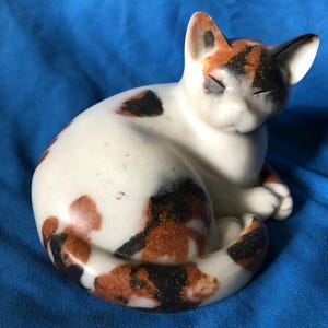 May include: A smooth, cream-coloured ceramic cat figurine in a curled, resting position. The cat features distinct patches of black and burnt orange throughout its body, including its head, back, and tail, with eyes depicted as closed slits.