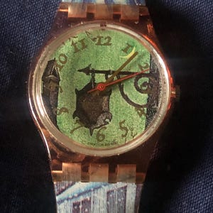 Puede incluir: Un reloj Swatch de oro rosa con una esfera verde y una ilustración en blanco y negro de una casa con una luna creciente. El reloj tiene una correa con un patrón blanco y azul.
