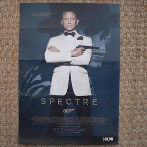 Puede incluir: Un póster de película para la película de James Bond Spectre, con Daniel Craig como James Bond en un esmoquin blanco, sosteniendo una pistola. El póster tiene un fondo azul con una imagen de calavera y el texto "SPECTRE 007" y "OCTOBER 26" en blanco. El póster también tiene el texto "ODEON" en blanco en la parte inferior.