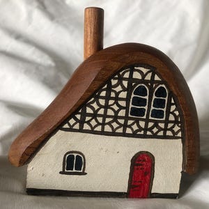 Può includere: Statuetta di cottage in legno con tetto di paglia marrone e camino. Il cottage ha una facciata bianca con elementi decorativi in bianco e nero, una porta rossa e infissi neri. Oggetto decorativo.