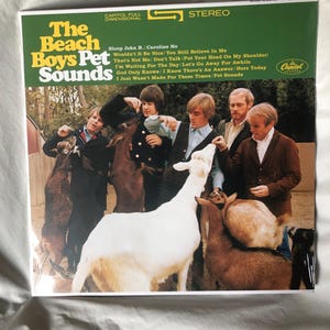 May include: The Beach Boys Pet Sounds album cover features a group of young men interacting with a white goat and other goats. The album title is printed in large, bold letters on a yellow background. The album's tracklist is printed below the title.