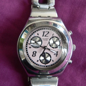May include: A silver Swatch watch with a pink face and black hands. The watch has a chronograph function and a date window.