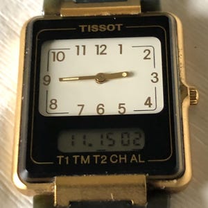 Op de afbeelding: Een Tissot horloge met een goudkleurige rechthoekige kast en een zwarte wijzerplaat. Het horloge heeft een witte analoge display met zwarte cijfers en wijzers, en een digitale display met de tijd. De horlogeband is donkergroen.
