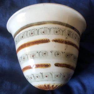 May include: A white ceramic planter with brown and gray painted stripes and floral designs. The planter has a wide opening and a rounded bottom.