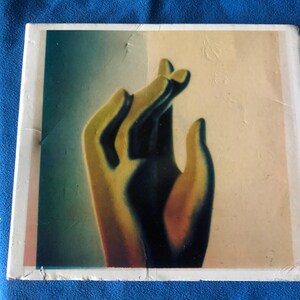 May include: Square artwork featuring a stylized hand in shades of yellow and blue. The hand is positioned against a backdrop of light colors, with a white border. The artwork has a vintage aesthetic.