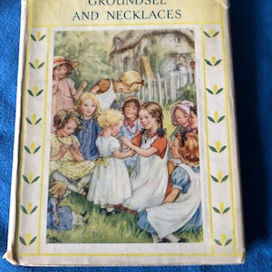 Puede incluir: Libro antiguo titulado "Groundsel and Necklaces" con una colorida ilustración de niños jugando al aire libre. La portada tiene un borde amarillo con detalles en verde. El libro está sobre un fondo azul.