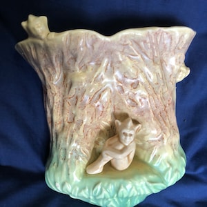 May include: A ceramic wall planter shaped like a tree trunk with a small gnome figure sitting on a ledge. The planter is glazed in shades of green, brown, and yellow.
