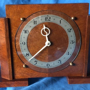 Puede incluir: Un reloj de madera vintage con una esfera de cristal transparente. El reloj tiene forma rectangular con un acabado marrón oscuro y una esfera circular con números y manecillas blancas. El reloj está sobre una superficie azul.
