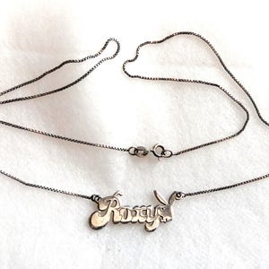 May include: A silver-toned necklace with the name "Roxy" in a cursive font, with a small bunny silhouette at the end. The necklace chain is a delicate box chain style, and the clasp is visible.