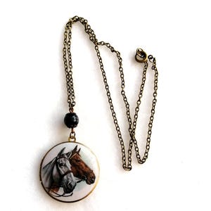 May include: A vintage-style necklace featuring a round pendant with a detailed painting of two horse heads. The pendant is suspended from a delicate, antique brass-toned chain. A dark bead accents the chain near the pendant.