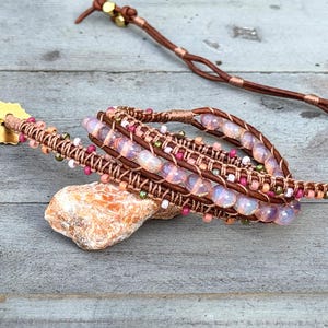 May include: A multi-strand wrap bracelet featuring brown leather cord, iridescent pale pink beads, and small accent beads in shades of pink, white, and metallic green. The bracelet includes a gold-toned star-shaped button closure and cord ends.